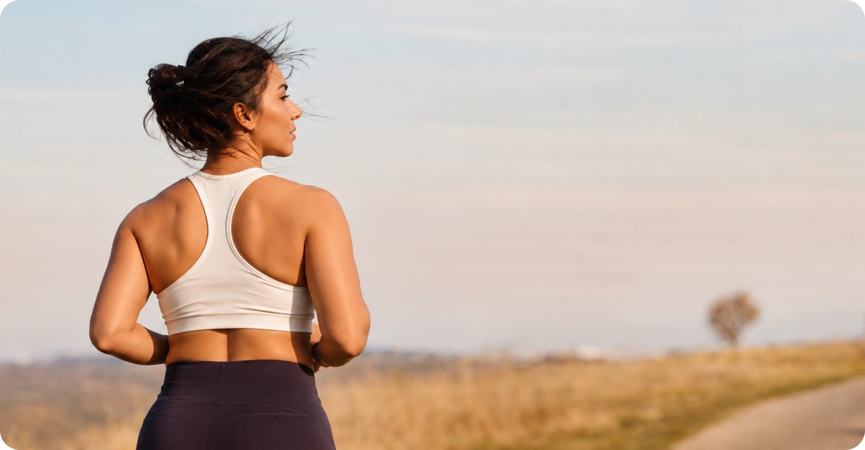 A woman in athletic wear seen from behind, looking out over a sunlit field under an open sky